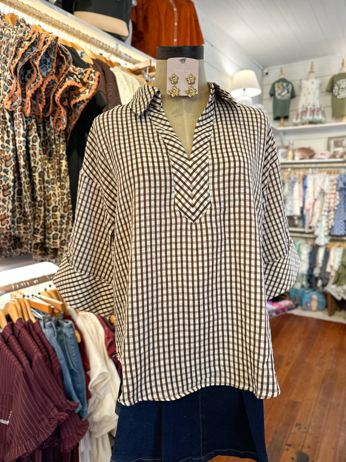 A brown and white gingham check long-sleeve blouse displayed on a dress form mannequin inside a clothing boutique, with racks of other garments visible in the background.