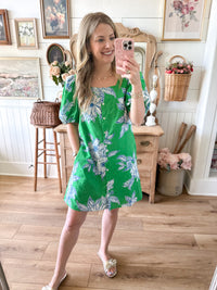 Woman taking a mirror selfie wearing a green floral dress in a home setting.