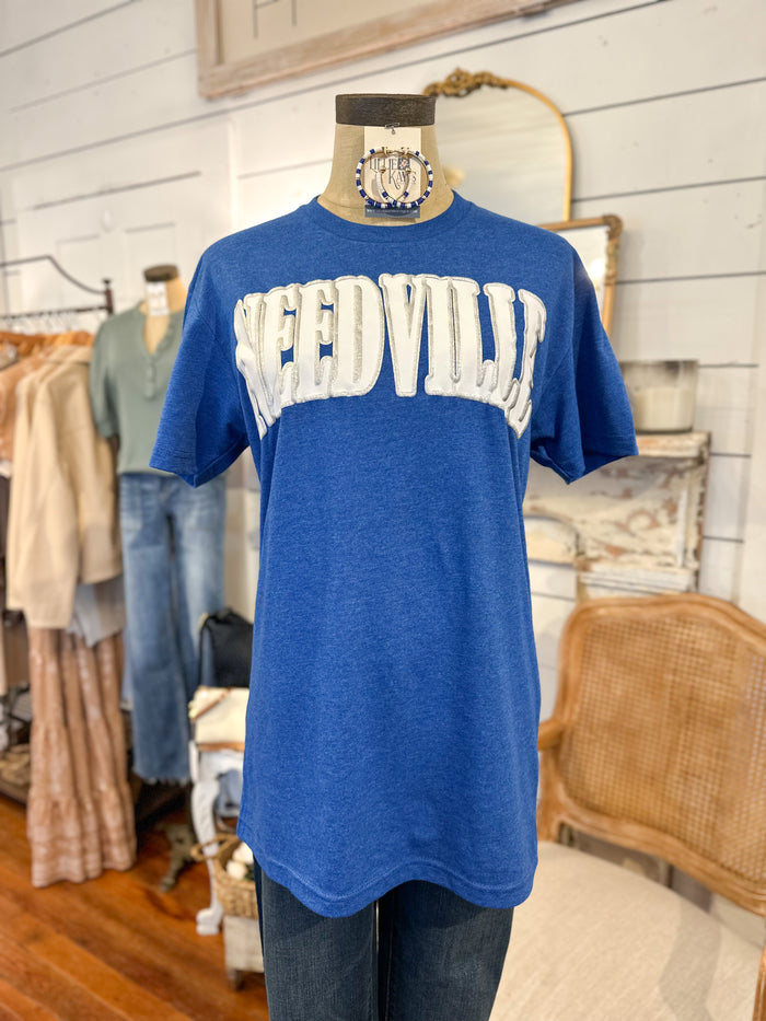 Blue t-shirt with 'NEEDVILLE' printed on it in a clothing store setting.