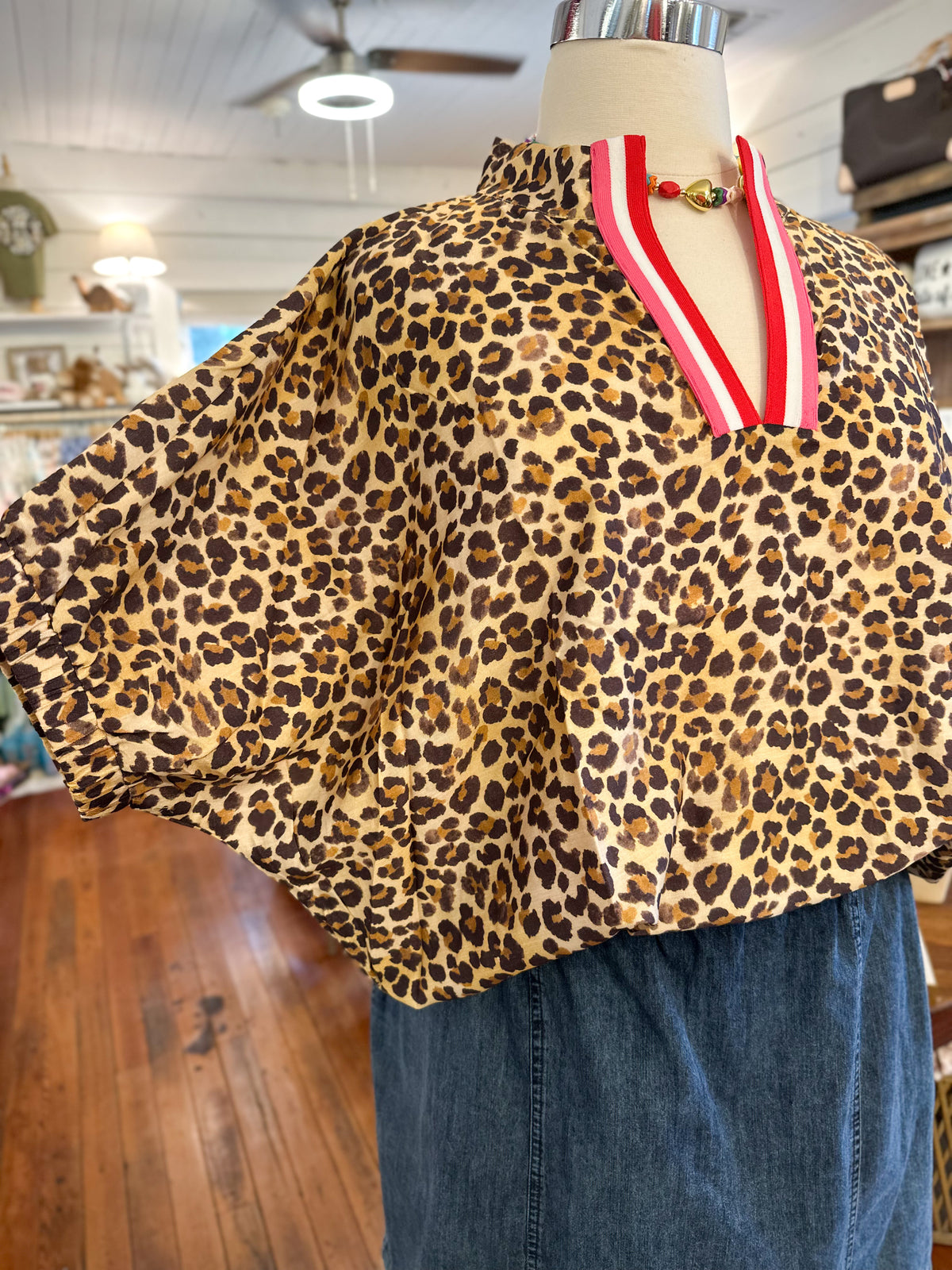 A leopard print blouse displayed on a mannequin torso in a retail store. The blouse has a beige base with brown and black leopard spots and a red-and-white striped V-neck trim.