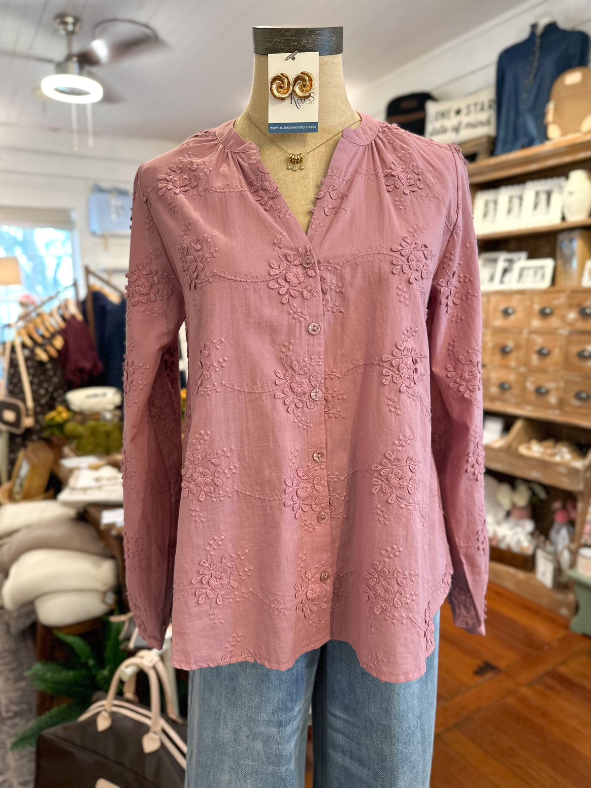 A dusty pink long-sleeve blouse with tonal floral embroidery, displayed on a mannequin torso in a clothing boutique. The blouse features a split neckline, button-front closure, and a relaxed fit, styled with light-wash blue jeans.