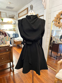 A black sleeveless dress with a high draped neckline, puffed shoulders, and a matching waist tie, displayed on a dress form inside a boutique.