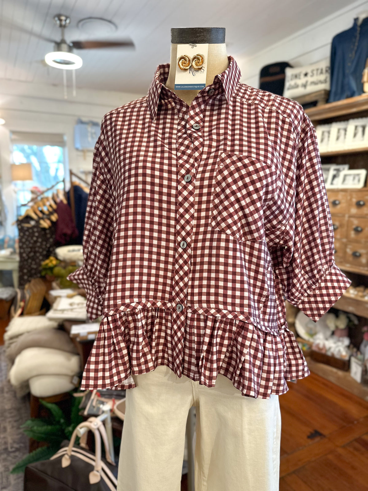 A maroon and white gingham button-up shirt with a ruffled hem, displayed on a mannequin in a boutique setting.