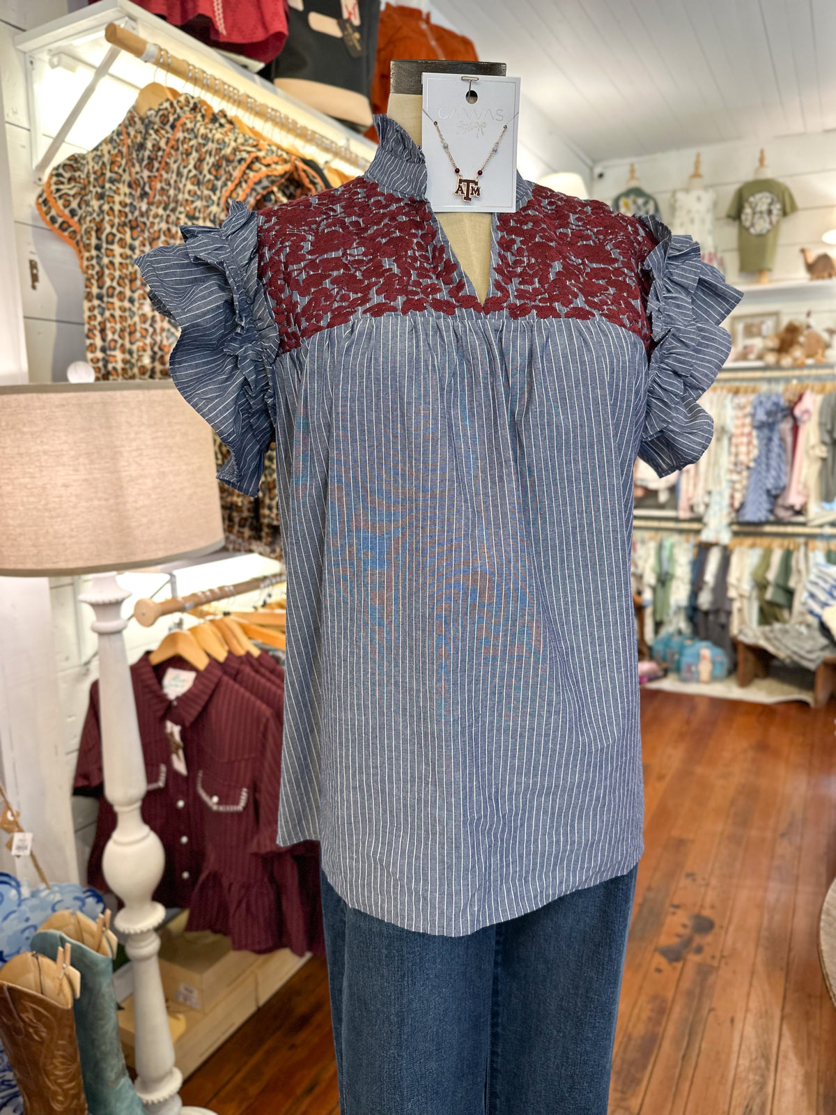 A women's blouse displayed on a mannequin in a clothing boutique. The top has a vertical blue-and-white stripe body,  burgundy floral embroidery across the chest, and ruffled sleeves.