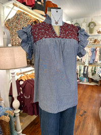 A women's blouse displayed on a mannequin in a clothing boutique. The top has a vertical blue-and-white stripe body,  burgundy floral embroidery across the chest, and ruffled sleeves.