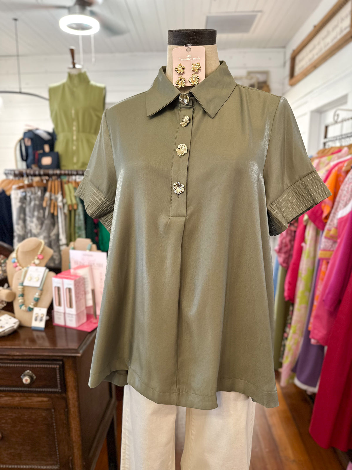 An olive green short-sleeve collared blouse with decorative gold-tone buttons, displayed on a mannequin torso inside a clothing boutique.