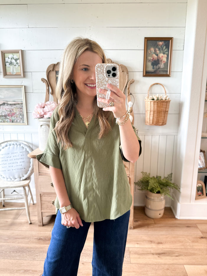 Woman taking a mirror selfie wearing an olive green top and blue jeans in a home setting.