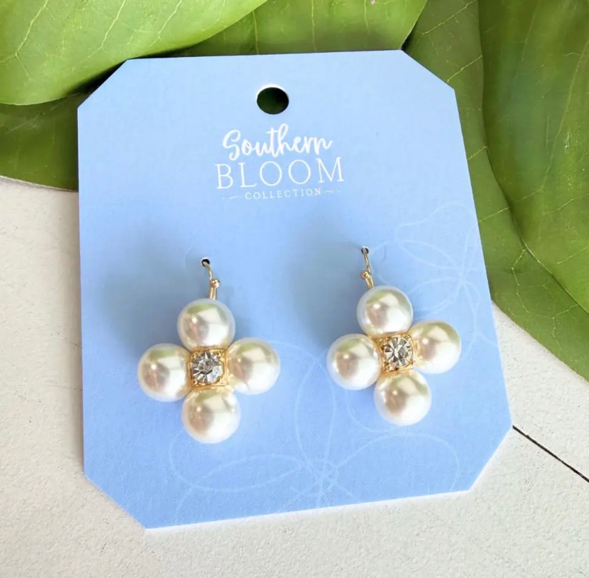 A pair of flower-shaped pearl earrings with gold-tone settings and a small clear crystal at the center, displayed on a light blue card labeled Southern Bloom Collection, with green leaves in the background.