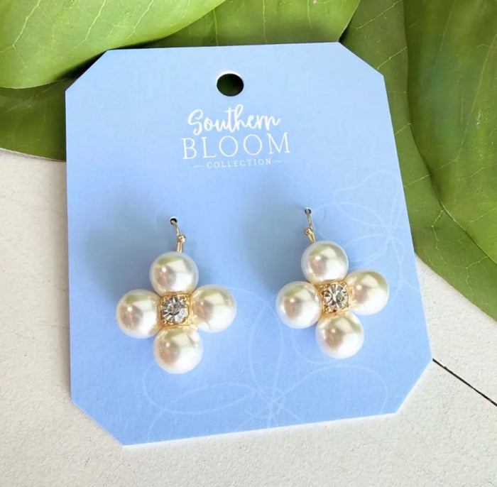 A pair of flower-shaped pearl earrings with gold-tone settings and a small clear crystal at the center, displayed on a light blue card labeled Southern Bloom Collection, with green leaves in the background.