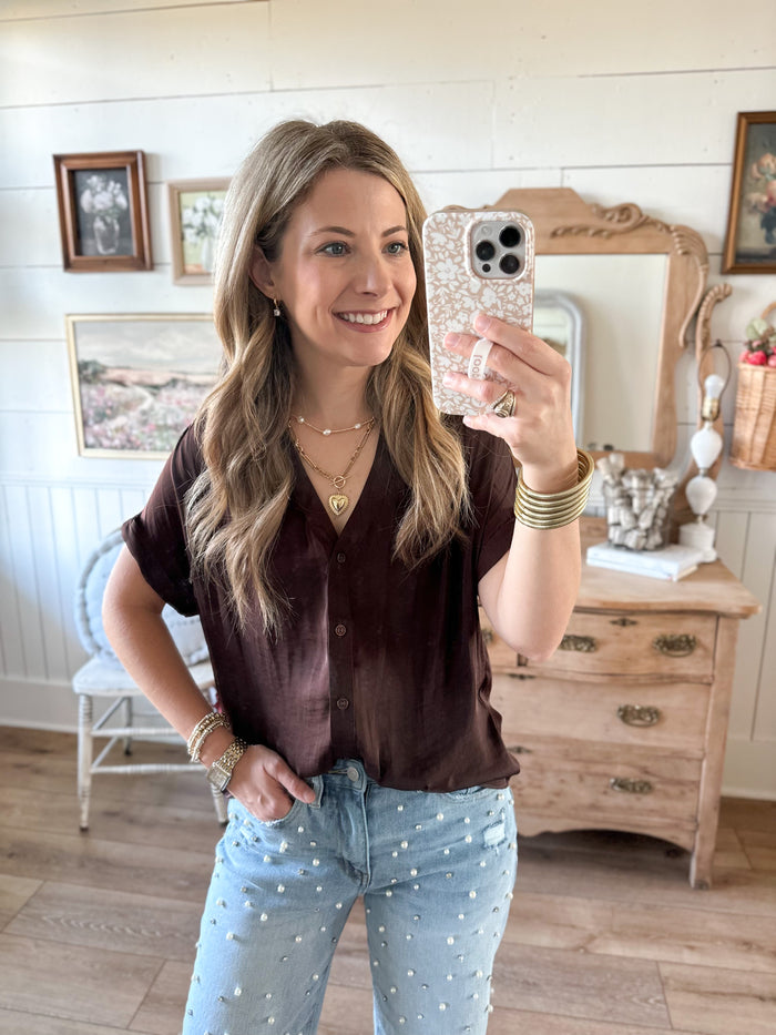 Woman taking a mirror selfie in a room with wooden floors and furniture. Brown silky soft button down top with rolled sleeves 