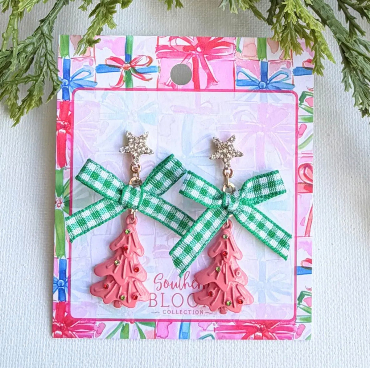 A pair of Christmas-themed dangle earrings displayed on their branded backing card. The earrings feature a green and white gingham ribbon bow with a gold-tone star charm above a pink plastic Christmas tree charm.