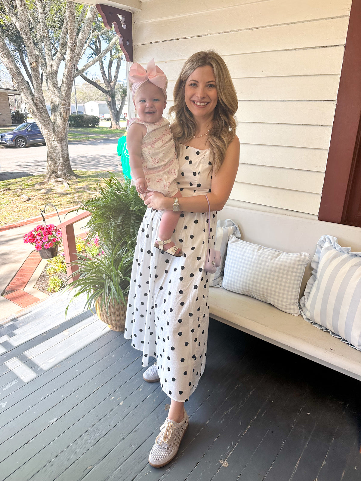 A woman stands on a porch holding a baby girl. The woman is wearing a white sleeveless maxi dress with black polka dots and white slip-on shoes.