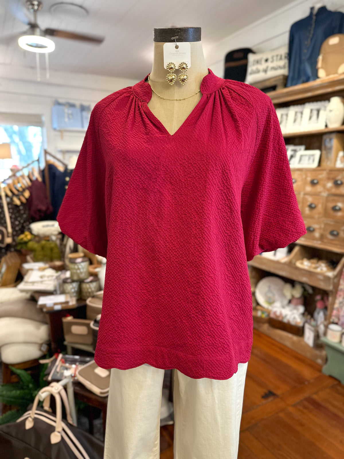 A woman's short-sleeve blouse in textured  fall red displayed on a mannequin torso inside a clothing boutique. The blouse features a gathered V-neckline and a scalloped hem.