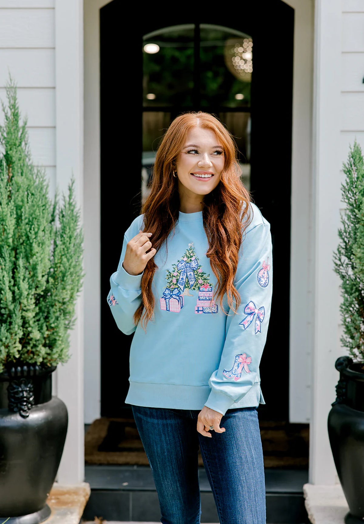 Woman wearing a light blue sweatshirt with colorful designs standing in front of a house entrance.