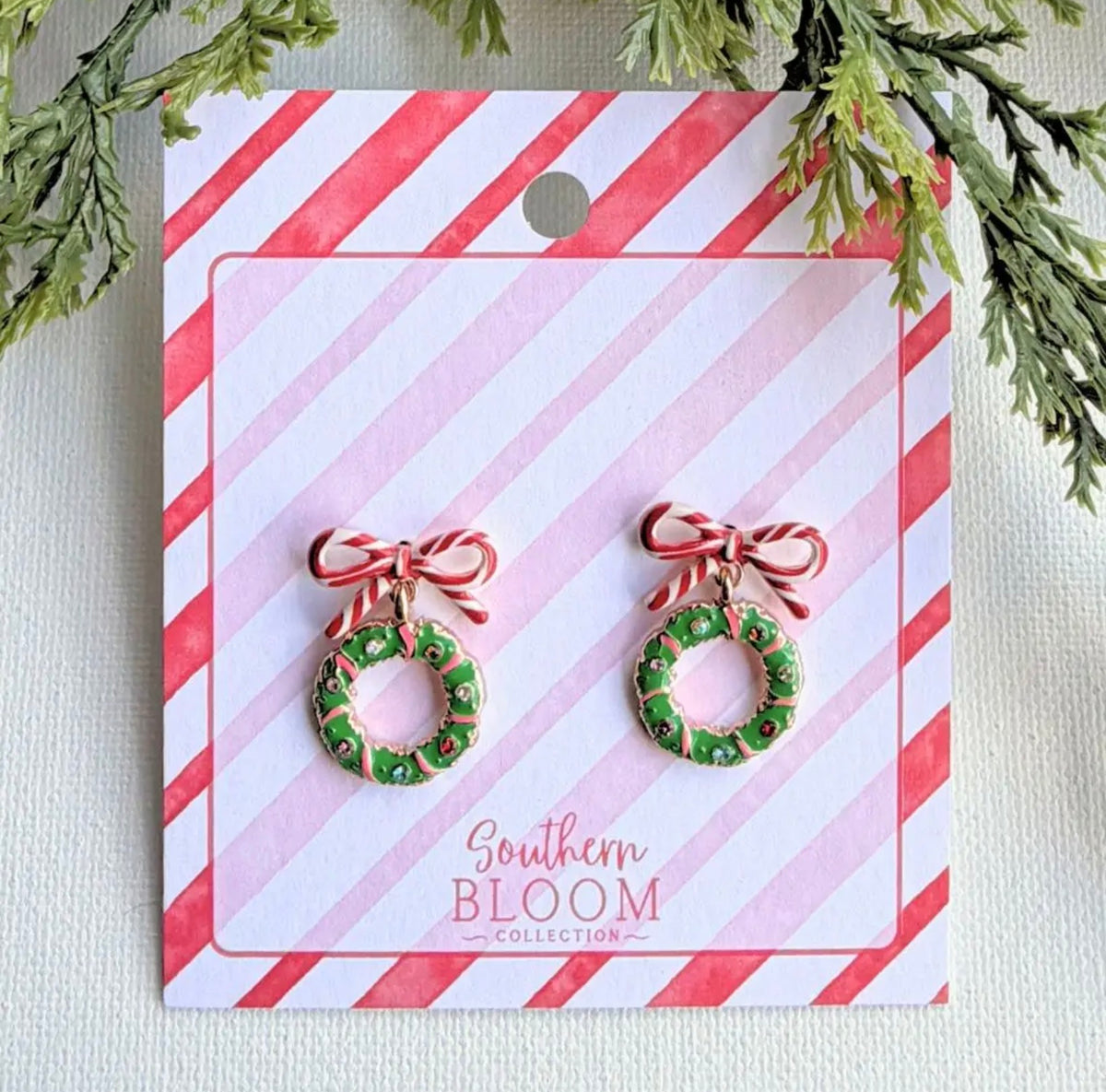 A pair of Christmas wreath stud earrings with red bows, displayed on a pink and white striped card labeled 'Southern Bloom Collection'. The card is styled with green pine branches at the top.