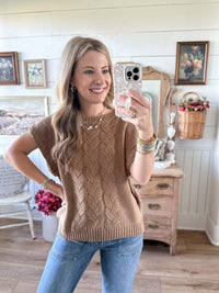 Woman taking a mirror selfie wearing a brown knitted sleeveless sweater and blue jeans in a home setting.