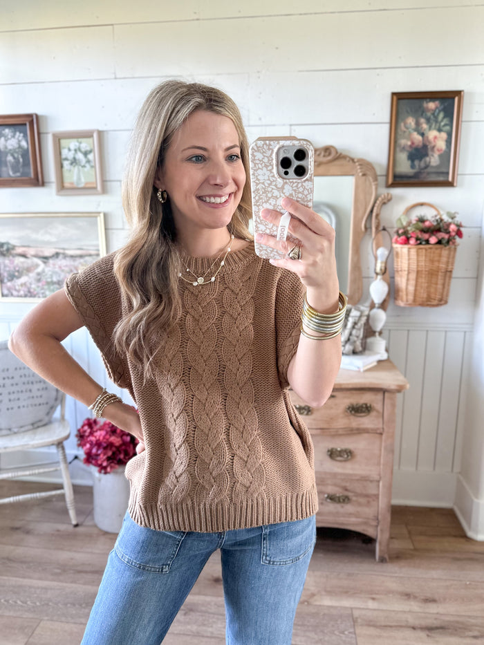 Woman taking a mirror selfie wearing a brown knitted sleeveless sweater and blue jeans in a home setting.