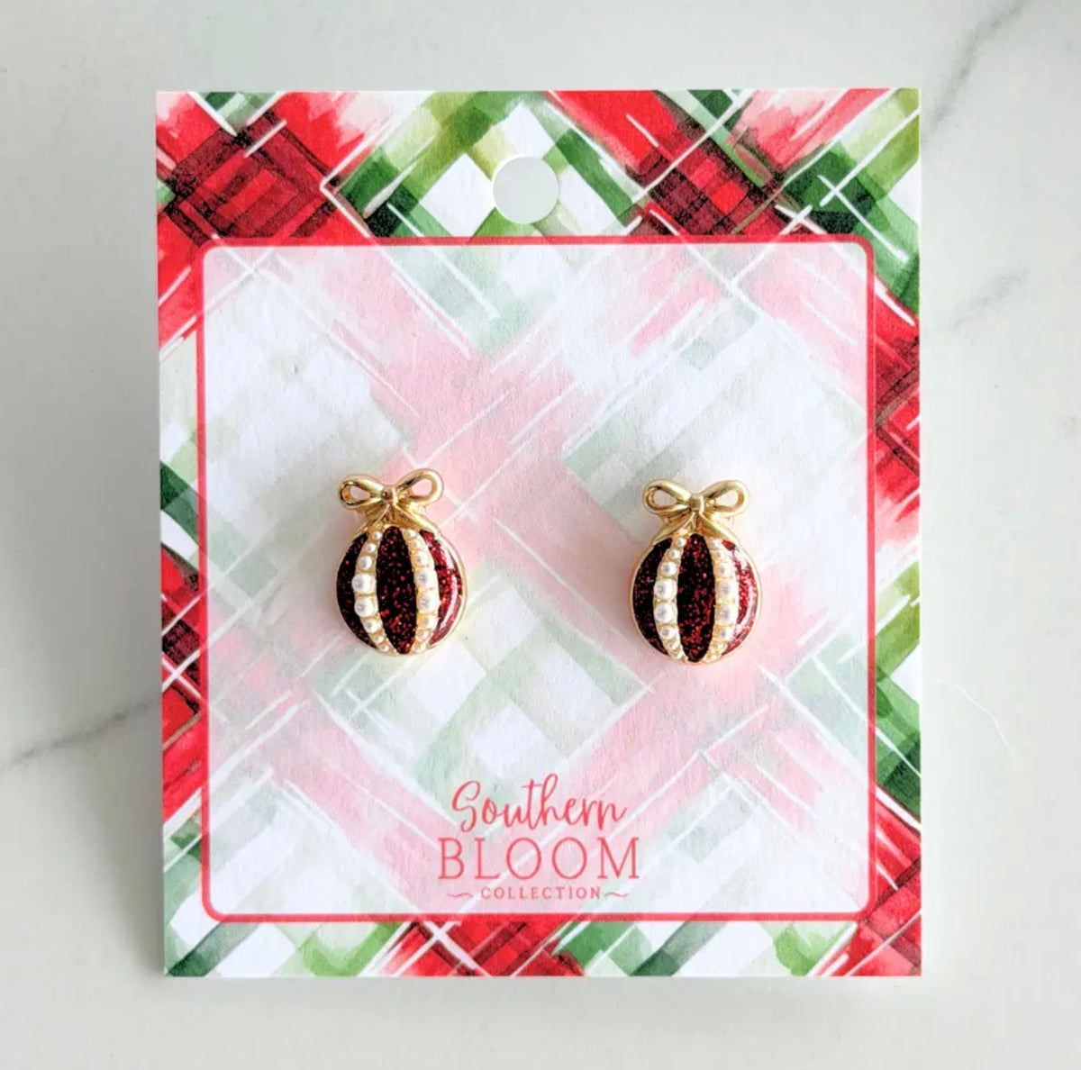 A pair of Christmas ornament stud earrings by Southern Bloom Collection, featuring red and white glittered ornaments with gold-tone borders and bows, displayed on a festive red, green, and white plaid backing card.