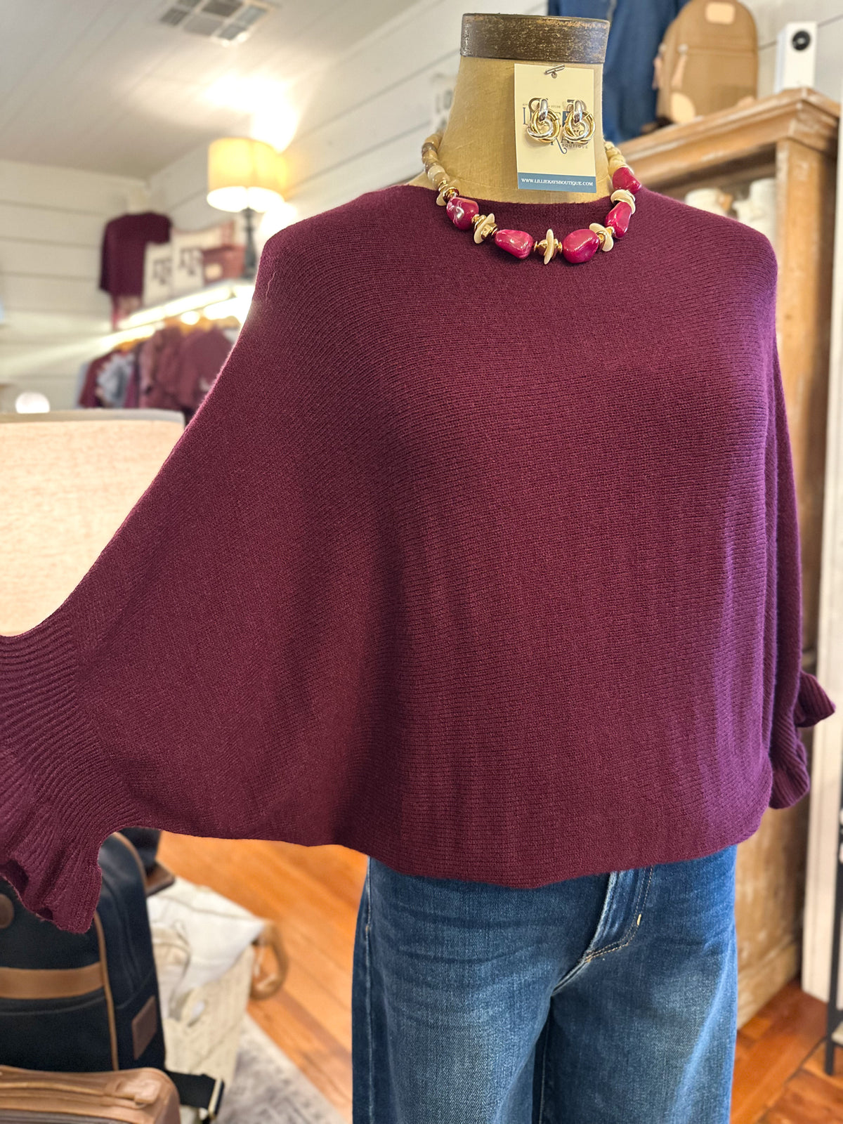 A burgundy knit poncho sweater displayed on a mannequin torso with blue jeans and a pink beaded necklace, inside a clothing store.