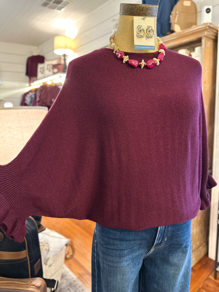 A burgundy knit poncho sweater displayed on a mannequin torso with blue jeans and a pink beaded necklace, inside a clothing store.