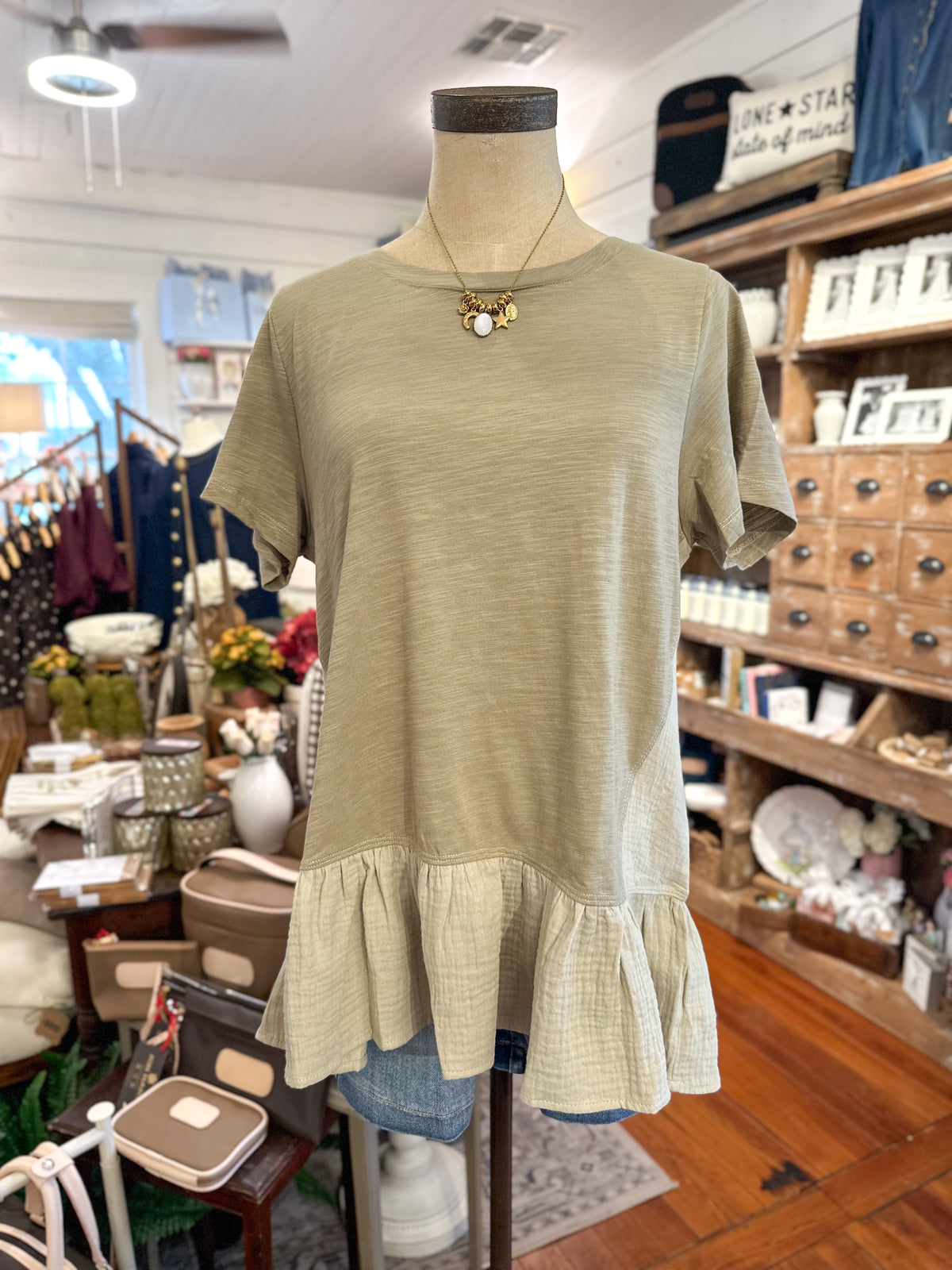 A short-sleeve top in a light olive color with a ruffled hem, displayed on a mannequin torso inside a boutique store.