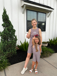 A woman and a young girl standing outdoors in front of a white house, both wearing matching sleeveless gingham dresses. The woman is taller and wears white sneakers, while the girl wears white sandals.