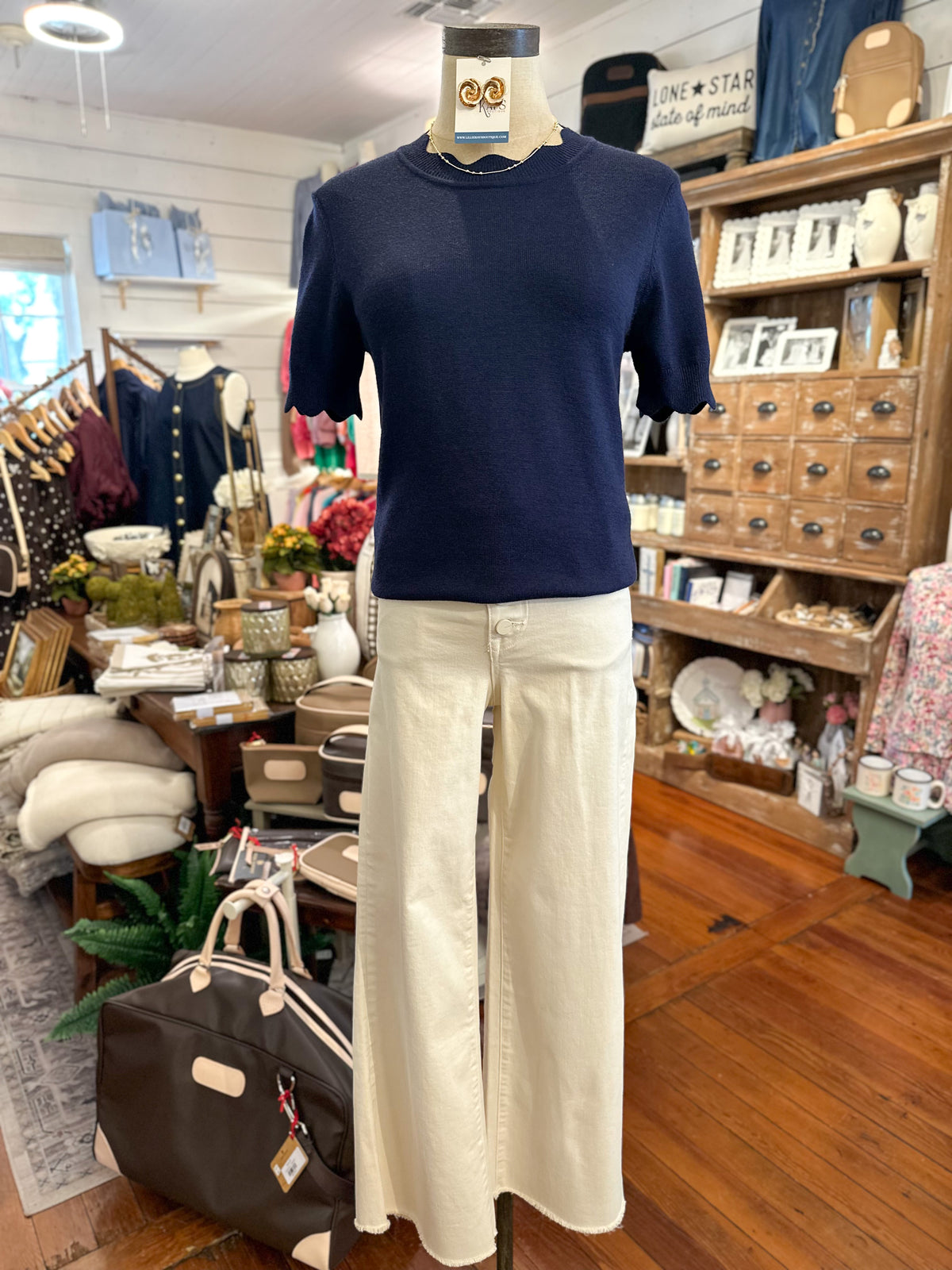 A navy blue short-sleeve knit top displayed on a mannequin torso, paired with off-white wide-leg pants, inside a retail boutique with wooden shelves and clothing racks in the background.