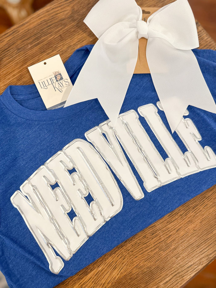 Blue t-shirt with white graphic and text on a wooden surface NEEDVILLE TEXAS 