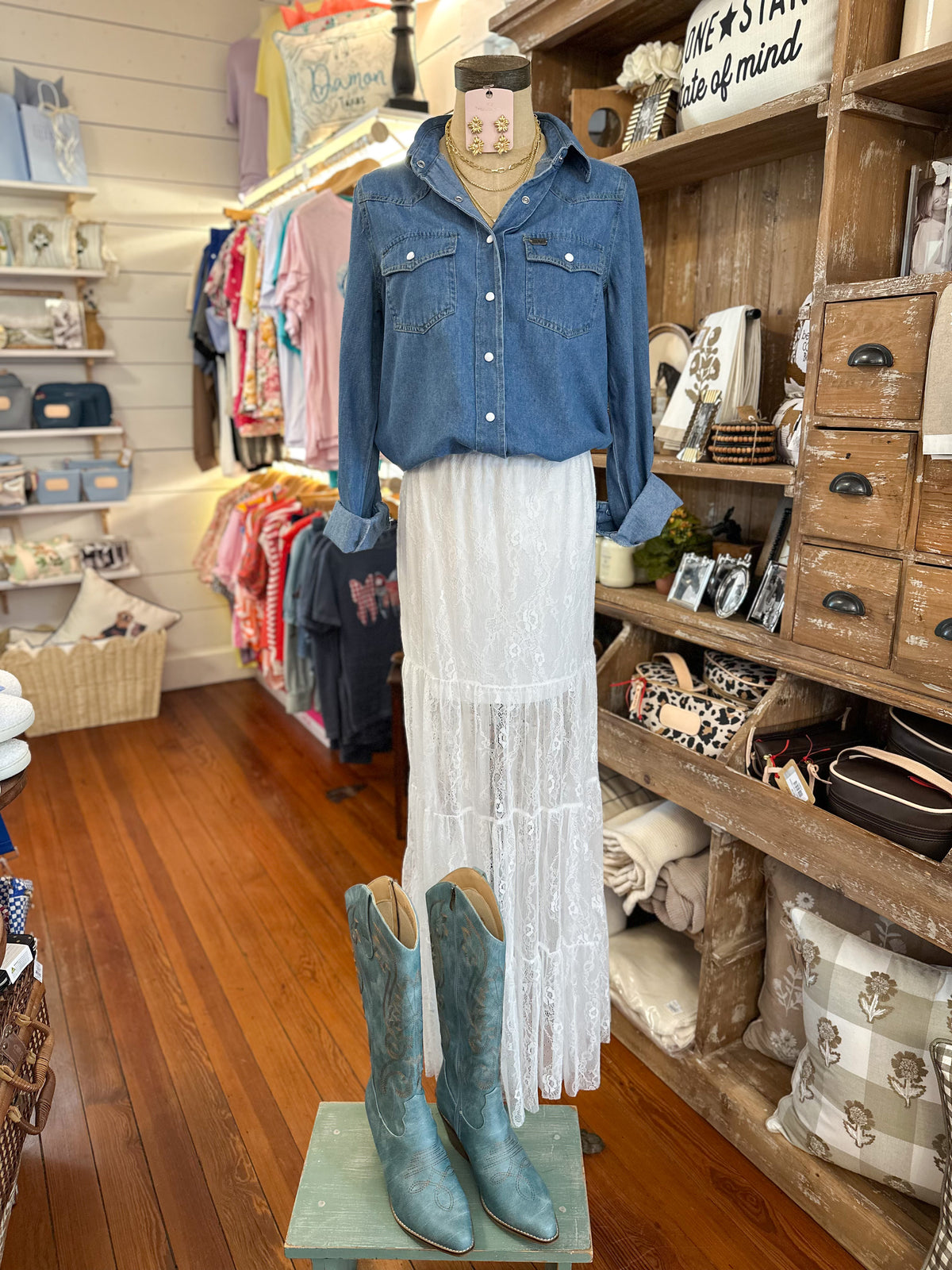 A mannequin in a rustic shop display wears a blue denim button-up shirt, a long white lace skirt, and blue cowboy boots on a wooden floor, with shelves of home goods and clothing in the background.