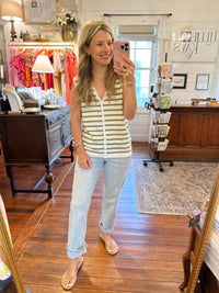 A woman takes a mirror selfie in a boutique while wearing a sleeveless white and olive green striped button-front top, light-wash cuffed jeans, and beige wedge sandals. The shop has wooden floors, a display cabinet, and clothing racks in the background.