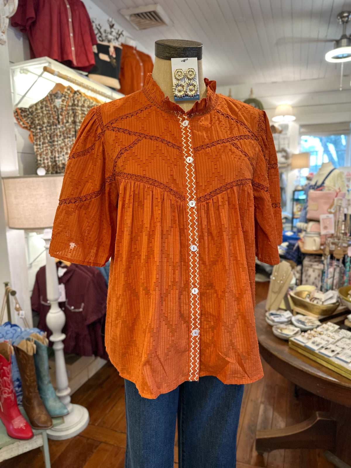An orange textured blouse with geometric embroidery and a decorative button placket, displayed on a mannequin torso inside a clothing boutique.