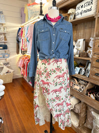 A two-piece women's outfit displayed on a mannequin in a boutique. The set includes a medium-wash blue denim button-up shirt tied at the waist and a cream-colored skirt with a pink horse and floral print.