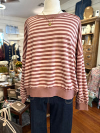 A long-sleeve striped top in dusty rose and cream is displayed on a mannequin torso inside a retail boutique. The top has horizontal stripes, a crew neckline, and ribbed cuffs and hem, paired with dark blue jeans on the mannequin.