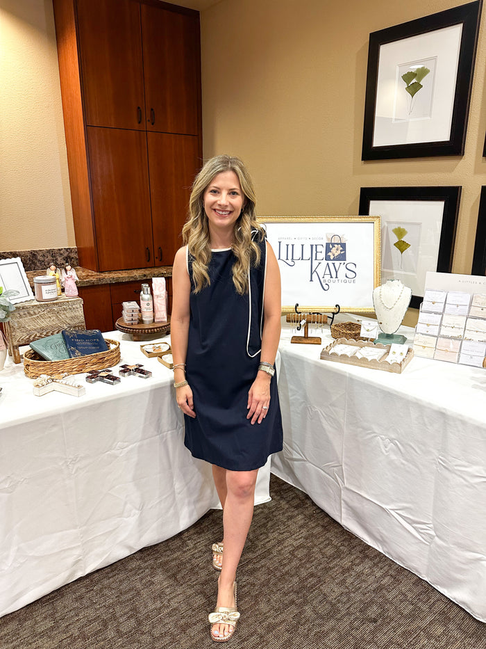 Woman in a navy dress standing in front of a table with Lillie Kay's products.