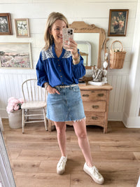 Woman taking a mirror selfie wearing a blue blouse with lace details and a denim skirt in a home setting.