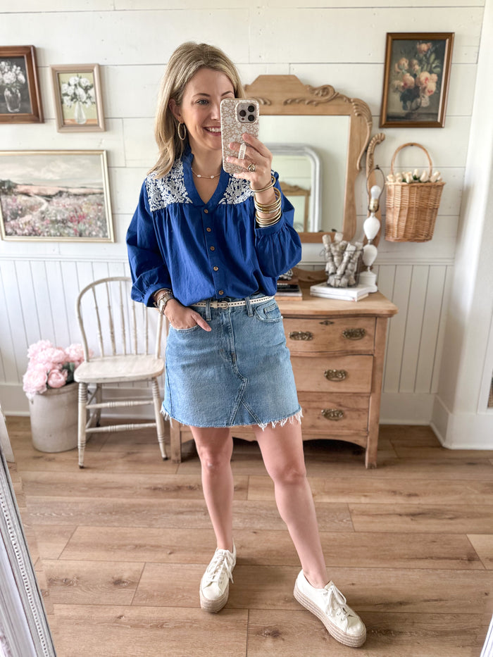 Woman taking a mirror selfie wearing a blue blouse with lace details and a denim skirt in a home setting.