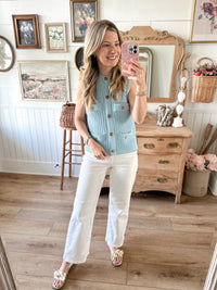 Woman taking a mirror selfie wearing a light blue sleeveless knit vest and white pants in a home setting.