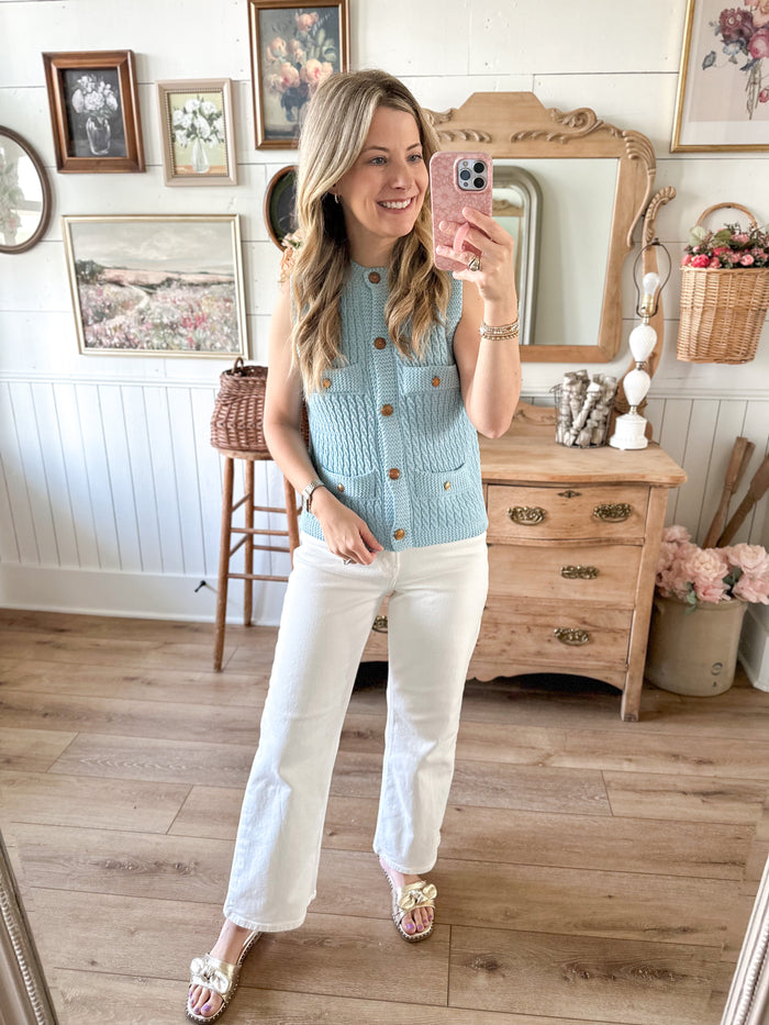 Woman taking a mirror selfie wearing a light blue sleeveless knit vest and white pants in a home setting.