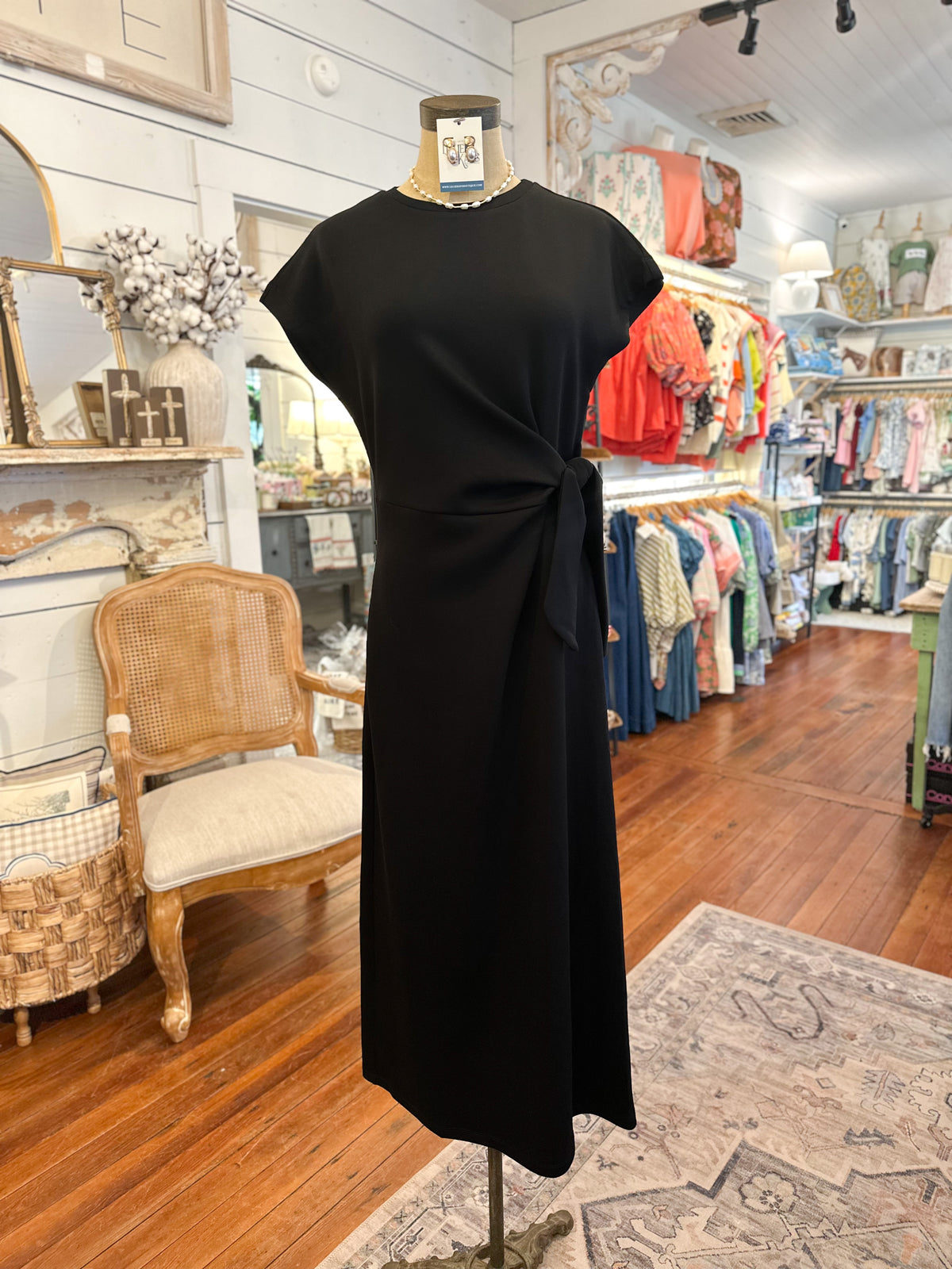 Black dress on a mannequin in a store setting with various clothing items in the background. Dress is soft material with a side tie 