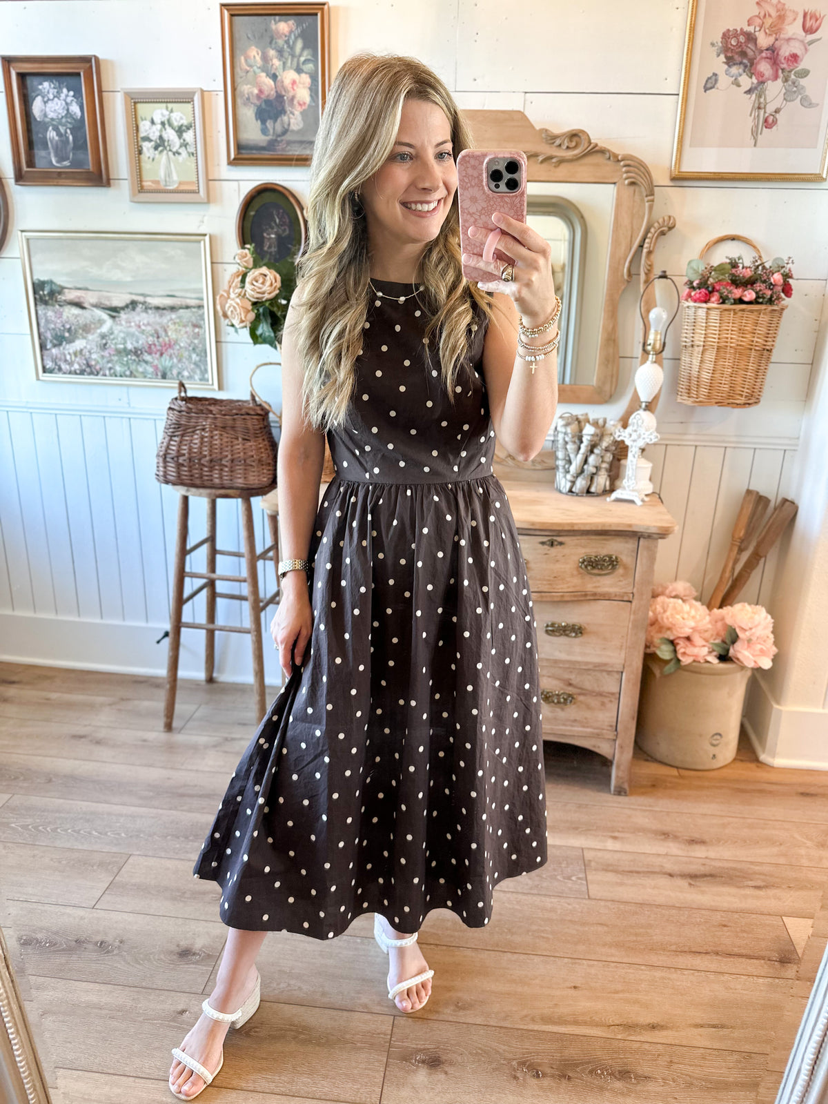 A woman takes a mirror selfie in a vintage-style room, wearing a sleeveless black midi dress with white polka dots and white heeled sandals.