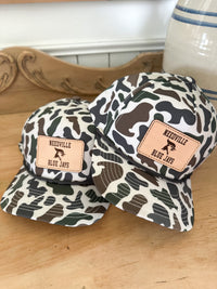 Two camouflage-patterned hats with leather patches on a wooden surface. Needville Blue Jays 