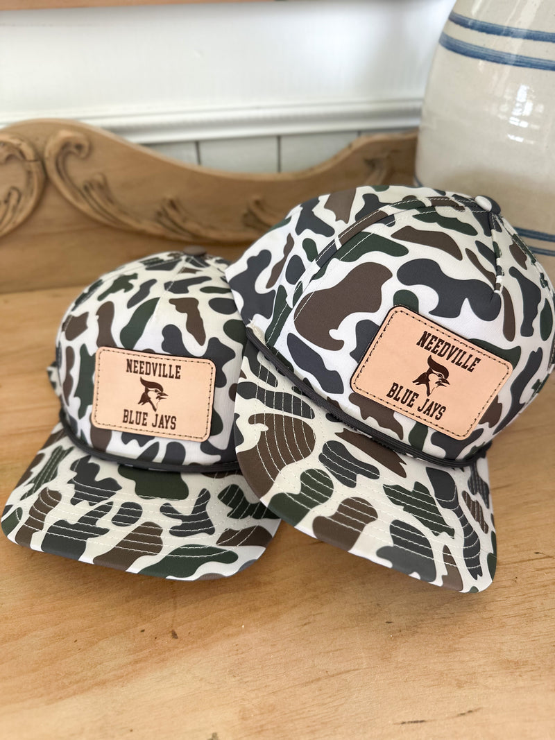 Two camouflage-patterned hats with leather patches on a wooden surface. Needville Blue Jays 