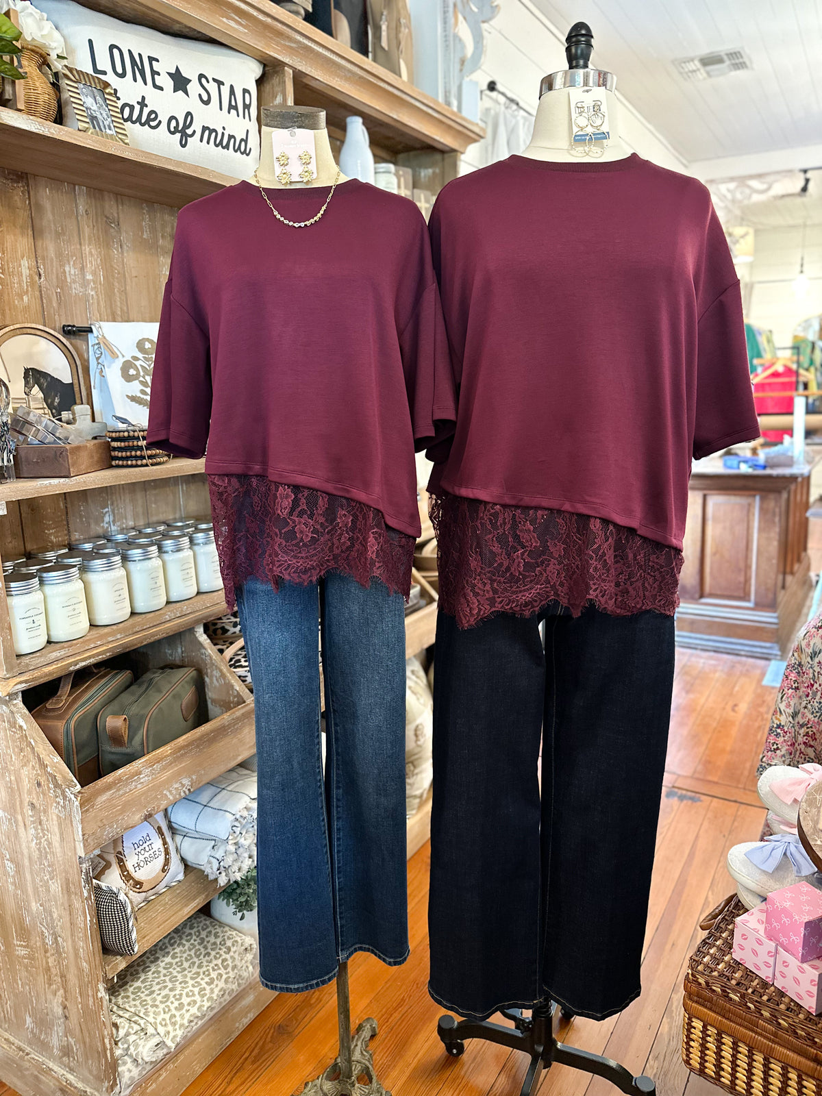 Two women's short-sleeve top dresses in deep burgundy are displayed on dress forms in a rustic boutique. The tops feature an asymmetrical hem with a sheer burgundy lace overlay.