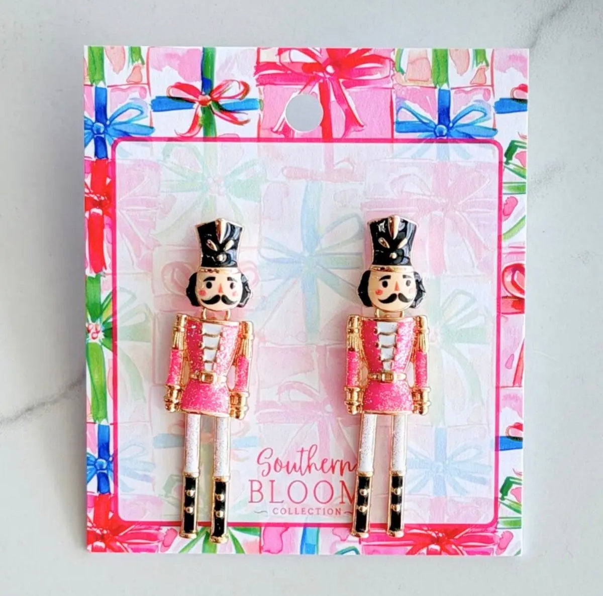 A pair of nutcracker stud earrings from the Southern Bloom Collection, displayed on their branded gift-wrapped card. The earrings feature pink and gold uniforms with black hats and boots, set against a light gray marble background.