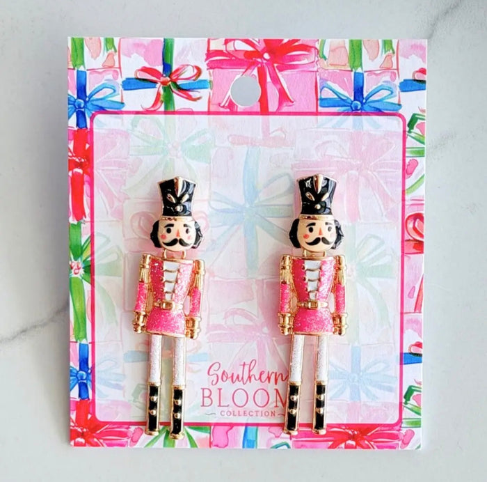 A pair of nutcracker stud earrings from the Southern Bloom Collection, displayed on their branded gift-wrapped card. The earrings feature pink and gold uniforms with black hats and boots, set against a light gray marble background.