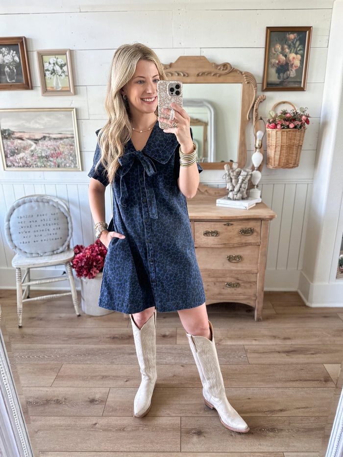 Woman taking a mirror selfie wearing a blue dress and white boots in a room with wooden flooring and furniture.