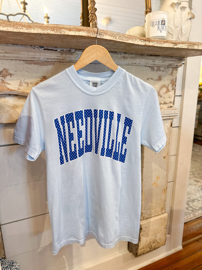 A light blue short-sleeve t-shirt hanging on a wooden hanger, featuring the word 'NEEDVILLE' printed across the chest in blue block letters with white polka dots. The shirt is displayed against a rustic, distressed wood fireplace mantel background.