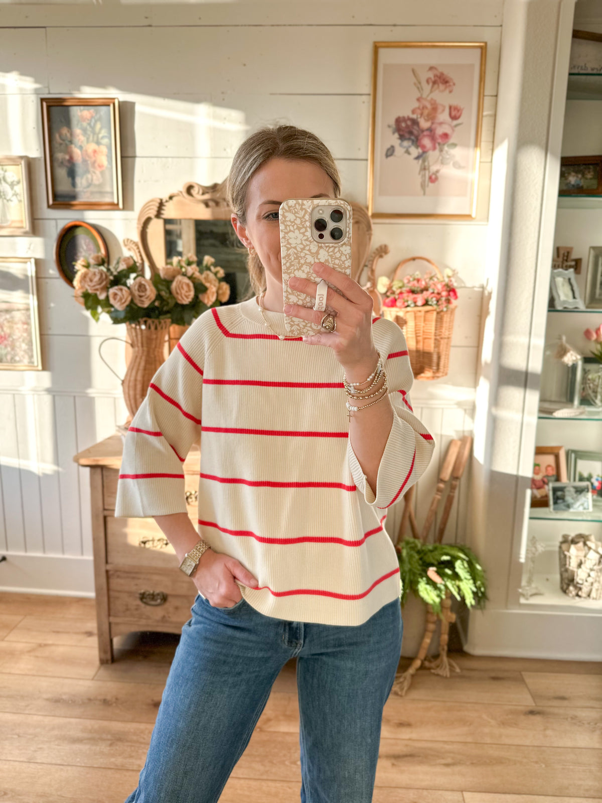 A woman takes a mirror selfie in a bright, sunlit room wearing a cream-colored short-sleeve top with thin red horizontal stripes and blue jeans.