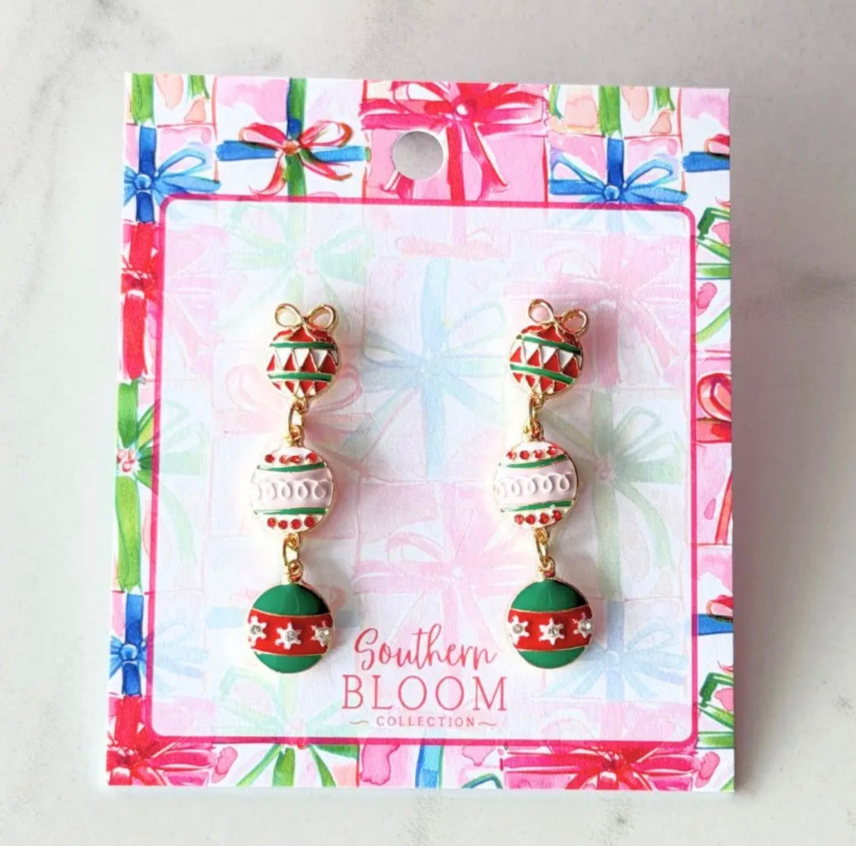 Christmas ornament dangle earrings by Southern Bloom Collection, displayed on their branded gift-wrapped card. The earrings feature gold-plated chains and beads decorated with red, green, and white holiday patterns.