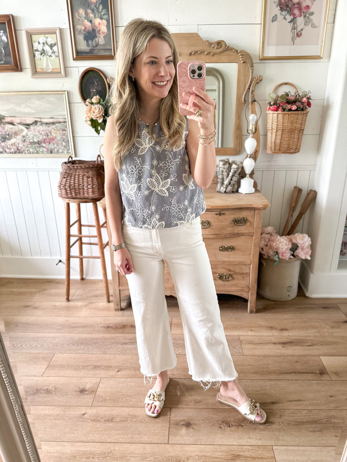 Woman taking a mirror selfie in a room with wooden floor and decor. embroidered top with black checkered print 